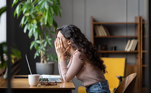 Woman sits in front of laptop covering her eyes with her hands