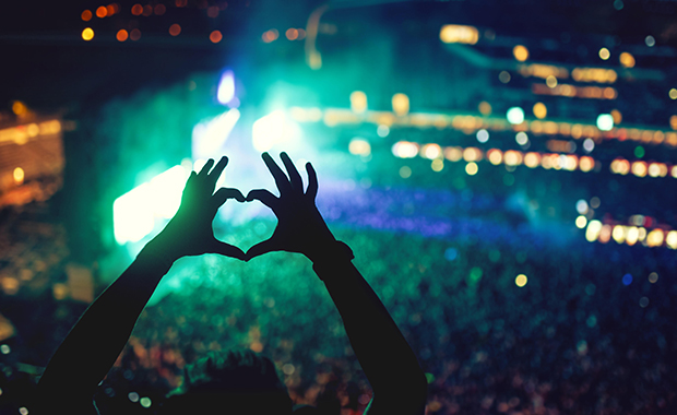 Hands held up in heart shape as silhouette in front of blur of lights in stadium concert