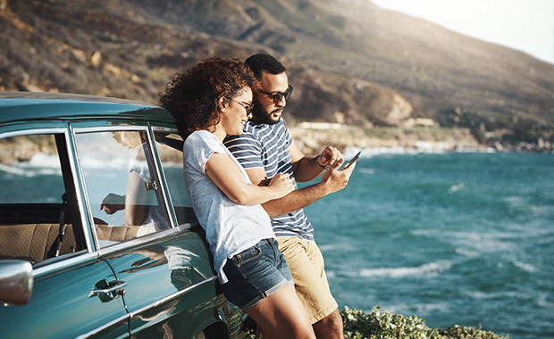 Man and woman lean against car on shore looking at phone