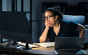 A woman sitting at a desk looks concerned as she stares at her computer screen with one hand covering her mouth.