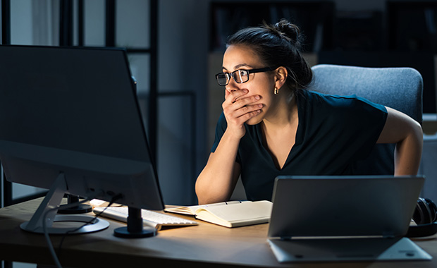 A woman sitting at a desk looks concerned as she stares at her computer screen with one hand covering her mouth.