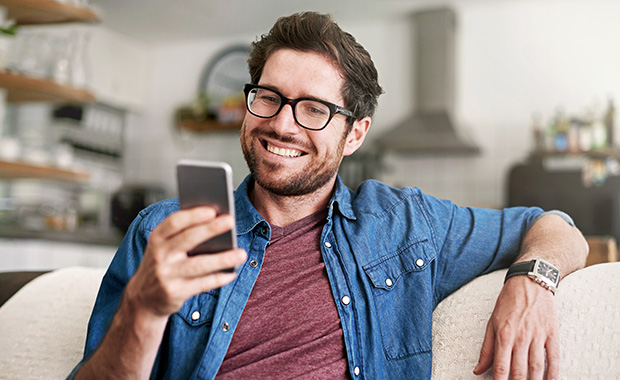 Smiling man with beard sits on couch looking at smartphone