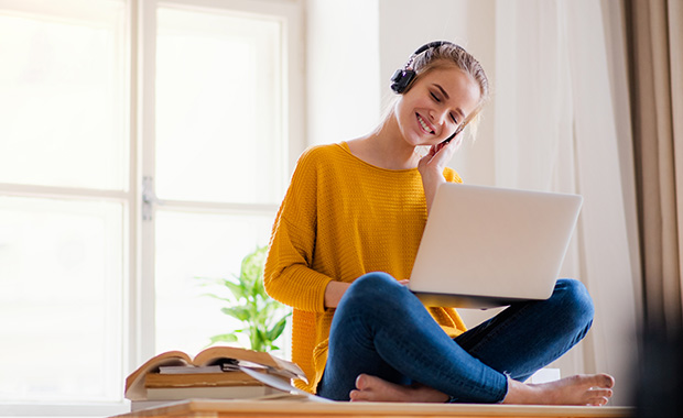 Student wearing headphones sits cross-legged with laptop open on her lap and a pile of books next to her