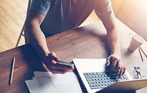 Hand holds a smartphone over a pile of papers on a desk while the other hand rests on a laptop keyboard