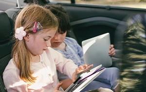Two young children sitting in the back seat of a car using tablets, focused on their screens.