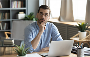 Man sits at desk in front of laptop with hand on chin