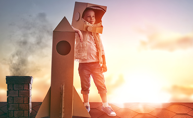 Child in astronaut costume stands on roof next to cardboard spaceship