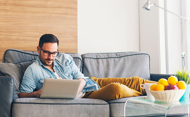 Man lying on brown couch looks at open laptop 