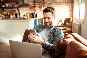 Man with beard on brown couch looking at smartphone and laptop