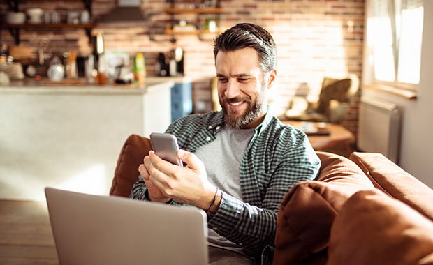 Man with beard on brown couch looking at smartphone and laptop