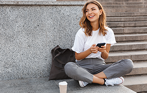 Smiling young woman sits cross-legged on ground holding smartphone