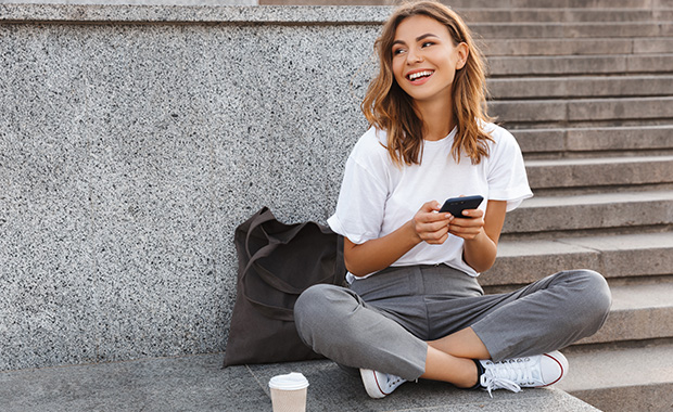 Smiling young woman sits cross-legged on ground holding smartphone