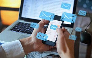 A person checking their smartphone for new emails while using a laptop, with digital envelope icons symbolizing incoming messages.