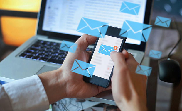 A person checking their smartphone for new emails while using a laptop, with digital envelope icons symbolizing incoming messages.