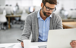 Man sitting at desk in office holding a pen and looking intently at laptop.