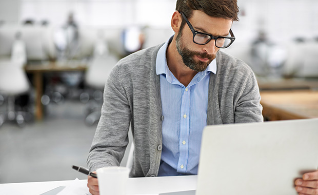 Man sitting at desk in office holding a pen and looking intently at laptop.