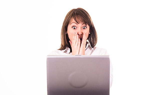 Young woman looks at laptop with shocked expression and hands to her mouth