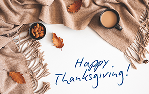 warm beige blanket, a cup of coffee, a small bowl of almonds, and scattered fall leaves with a handwritten text that reads Happy Thanksgiving!