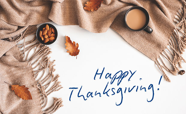 warm beige blanket, a cup of coffee, a small bowl of almonds, and scattered fall leaves with a handwritten text that reads Happy Thanksgiving!