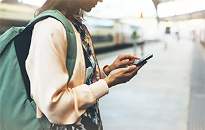 A person standing on a train platform checks their smartphone while wearing a backpack.