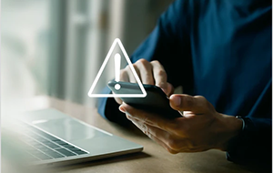 A person sitting at a desk using a smartphone next to an open laptop with a warning icon overlayed.