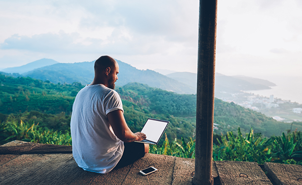 Man with laptop sits on wooden deck looking over mountains and shoreline