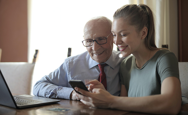 Smiling young woman and senior man sit in front of laptop looking at phone