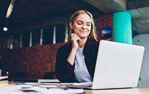 Smiling young woman looks at laptop on desk covered with papers