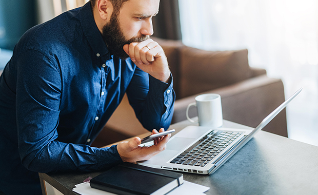 Frowning, bearded man with hand on chin holding smartphone and looking at laptop