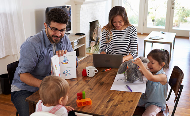 Family of four sits around table with father reading to toddlers and mother working on laptop