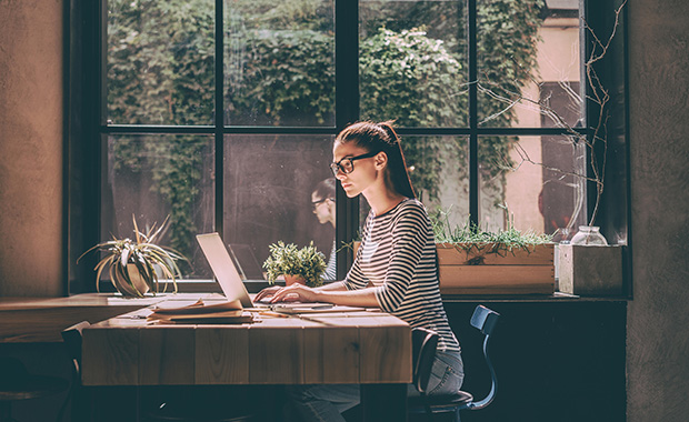 Young woman sits at desk in front of window working on laptop