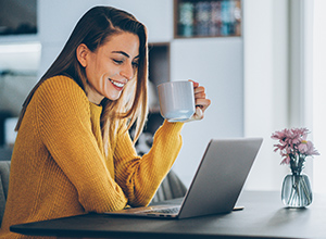 Woman wearing yellow sweater sits at kitchen table using laptop