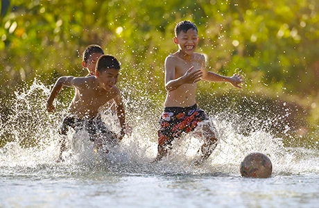 Drei Jungen in Badehosen rennen im Wasser lachend einem Ball hinterher.