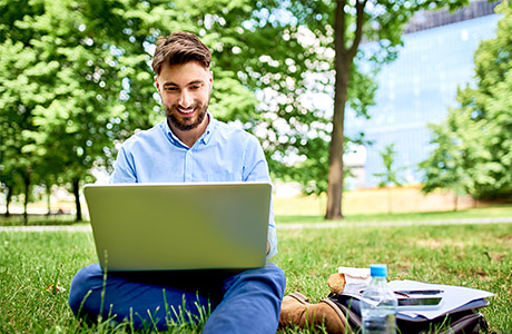 Junger Mann mit braunem Vollbart sitzt auf einer grünen Wiese mit einem Laptop auf dem Schoß und arbeitet