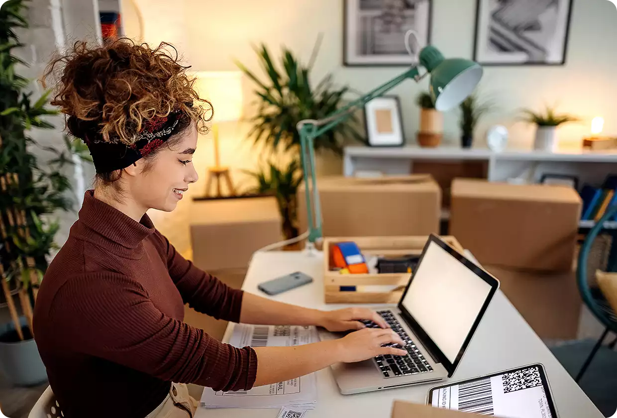 Frau sitzt am Schreibtisch vor einem Laptop und tippt. Im Hintergrund sind Umzugskartons zu sehen.