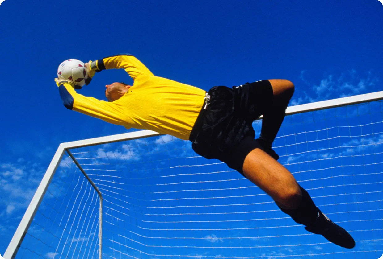 Fußballtorwart in gelbem Shirt vor blauem Himmel, der den ankommenden Ball auffängt.