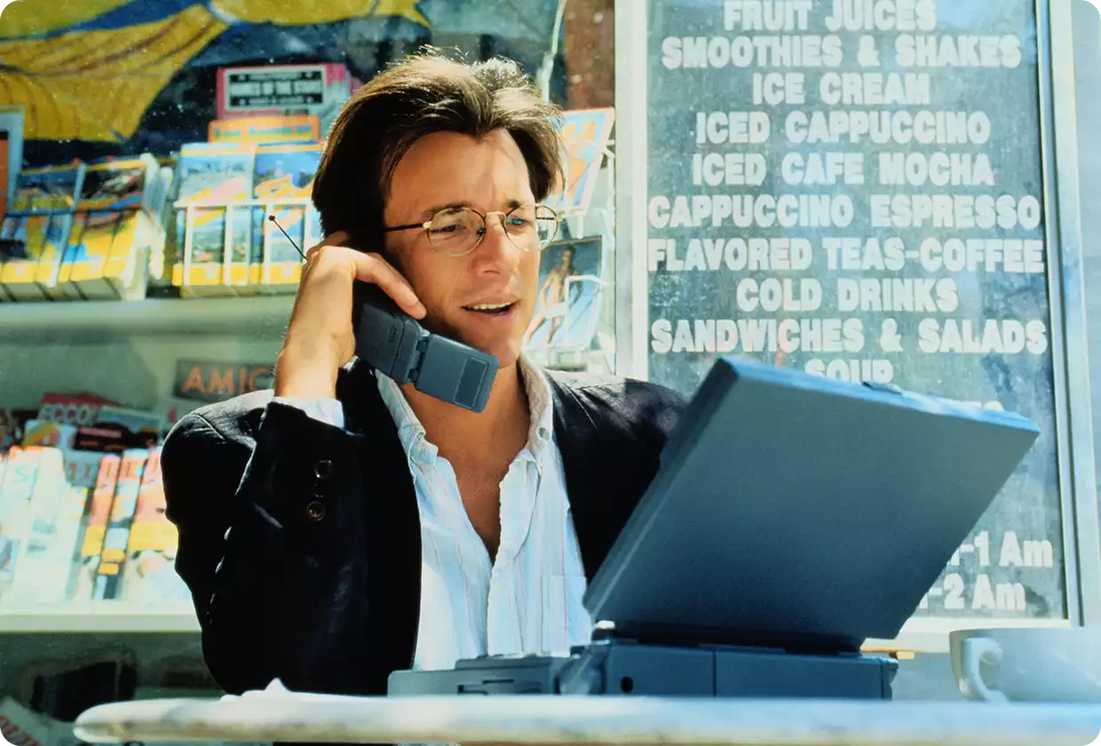 Mann hält ein Handy mit Antenne in der Hand und tippt auf seinem Laptop - Szene aus den 90ern.