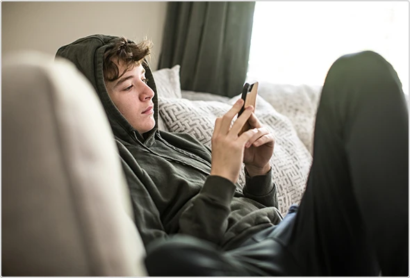 Junger Jugendlicher liegt auf Couch und schaut resginiert auf sein Smartphone