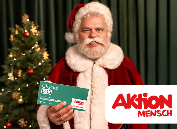 Person in rotem Weihnachtsmannkostüm hält ein grünes Los mit der Aufschrift 'Glücks LOS' vor einem geschmückten Weihnachtsbaum.