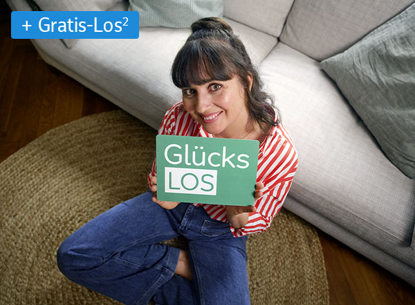 Frau mit rot-weiß gestreiftem Hemd hält rotes Smartphone vor sich, sitzt an Holztisch vor Bücherregal und Fenster mit Blick ins Grüne.