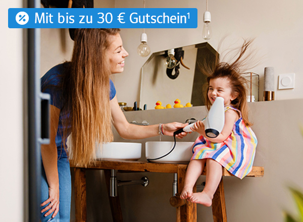 Eine Frau föhnt einem kleinen Mädchen in einem Badezimmer die Haare. Die Haare des Mädchens sind durch den Fön zerzaust. Beide lächeln fröhlich.