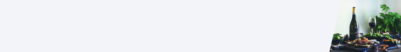 Flasche Rotwein mit goldener Kapsel und schwarzem Etikett 'Unico', daneben Weinglas mit Rotwein, grüne Trauben und Tannenzapfen auf Tisch.