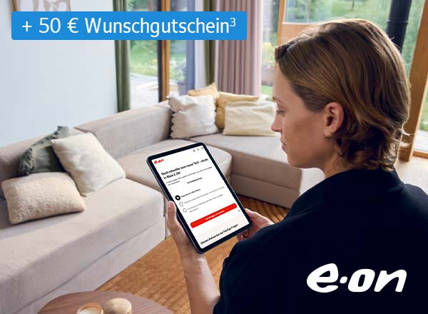 Person mit braunen Haaren hält Tablet mit E.ON-Webseite, sitzt vor hellgrauem Sofa mit Kissen in hellem Wohnzimmer.
