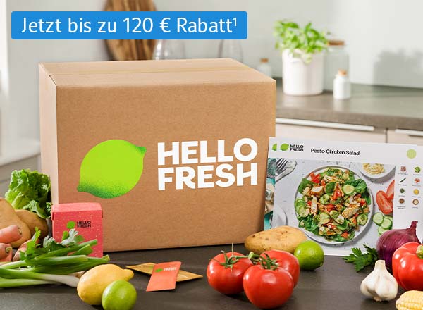 Eine HelloFresh Essensbox steht auf einem Tisch. Um die Box herum sind Kartoffeln, Paprika, Lauch, Mais, und weiteres Gemüse drapiert. An der Essensbox lehnt eine Rezeptkarte mit einem Kochrezept. Im Hintergrund sind die Umrisse einer Küchenzeile erkennbar.