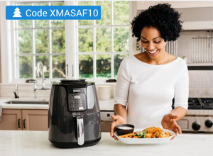 Frau in weißem Oberteil steht in moderner Küche vor schwarzer Heißluftfritteuse und hält Teller mit frittiertem Essen und Dip-Schale.