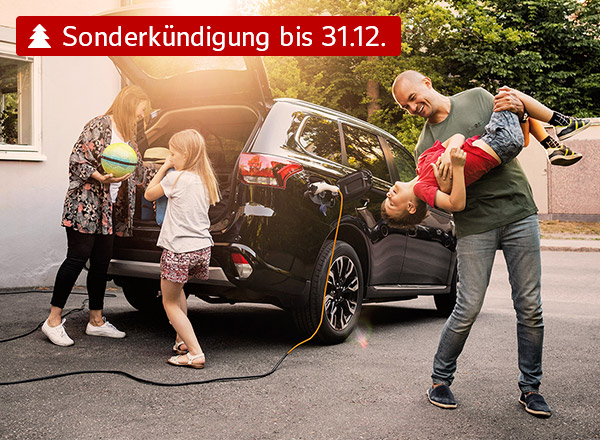 Ein Auto mit geöffnetem Kofferraum steht vor einem Haus. Eine Familie steht vor dem Auto. Die Mutter lädt Gepäck in den Kofferraum, ein Kind steht daneben. Der Vater steht scherzend mit einem weiteren Kind neben dem Fahrzeug.