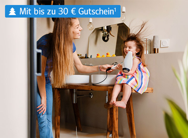 Eine Frau föhnt einem kleinen Mädchen in einem Badezimmer die Haare. Die Haare des Mädchens sind durch den Fön zerzaust. Beide lächeln fröhlich.