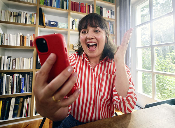 Person hält ein Smartphone in einem Raum mit Bücherregal, unten Logo ‚GlücksLOS‘.