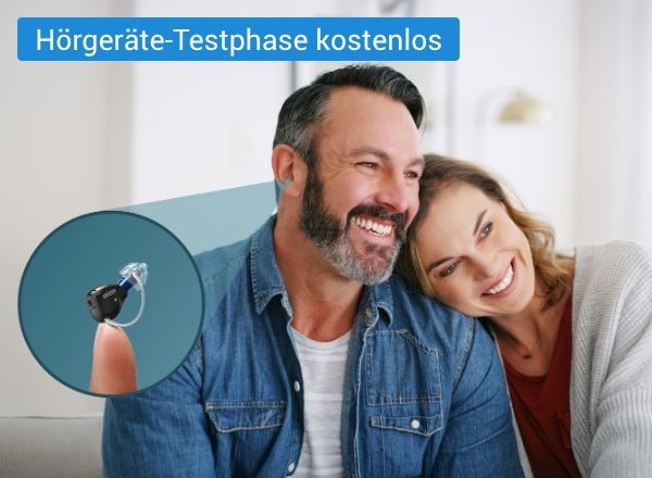 Ein lächelndes Paar sitzt eng nebeneinander in einem hellen Raum. Im Vordergrund ist zusätzlich ein kleines Hörgerät zu sehen, das auf einem Finger präsentiert wird.