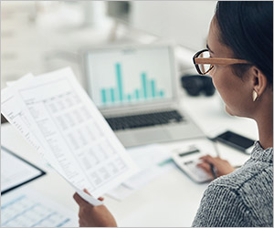 Eine Person analysiert Dokumente mit Laptop und Taschenrechner an einem Schreibtisch.